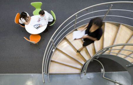 Lady walking down spiral stairs with students sat at the bottom of the stairs working collaboratively 
