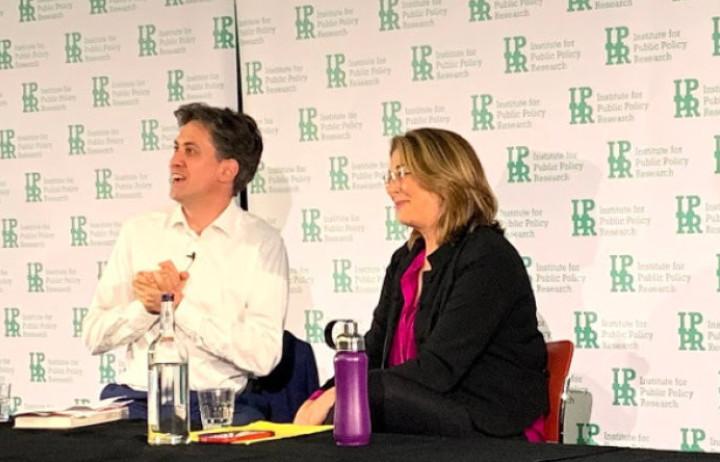 Ed Miliband and Naomi Klein speak to an audience in front of a white screen that reads IPPR (Institute for Public Policy Research). Ed wears a white shirt and Naomi wears a dark cardigan.