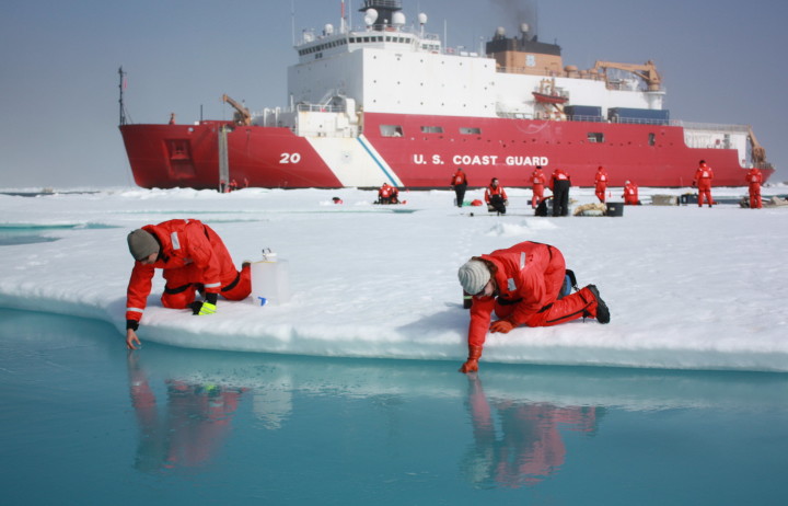 Photo of a person wrapped up in col-weather outfit crouching on the edge of a floating ice-sheet to collect a sample from a body of water.